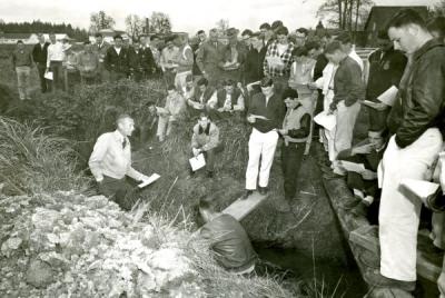 Art King, standing in ditch and wearing light colored jacket, addressing a group of students, 1967.